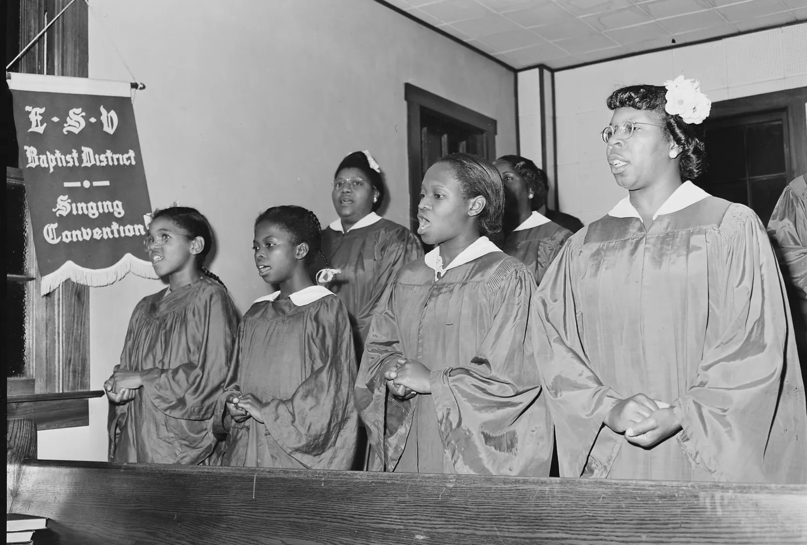 A black-and-white archive photo of a robed church choir of women and girls singing beside a Baptist District Singing Convention banner