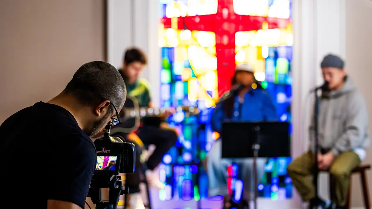 A videographer filming a three-person worship team on a camera in a church, with a stained glass cross behind the musicians