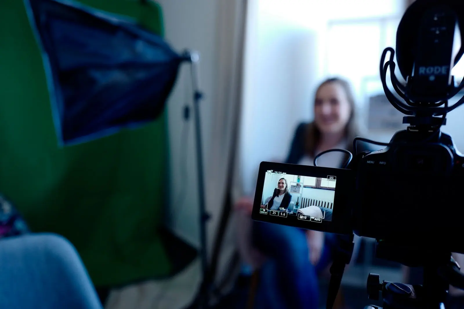 A camera's screen showing a smiling woman being interviewed, with a softbox light beside her and the woman out of focus behind the camera