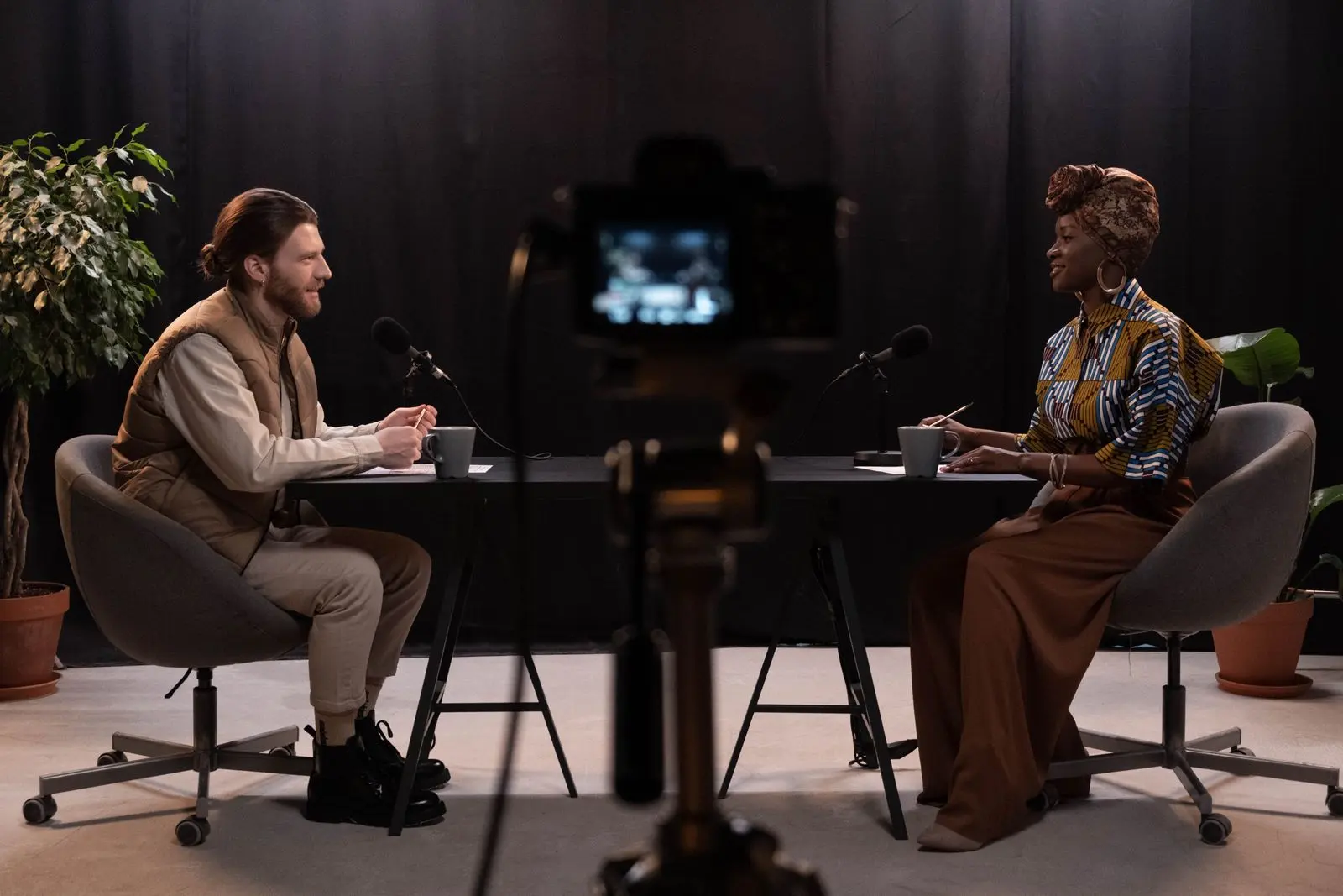 A man and a woman in a relaxed sit-down conversation at a table with microphones, filmed by a camera in the foreground
